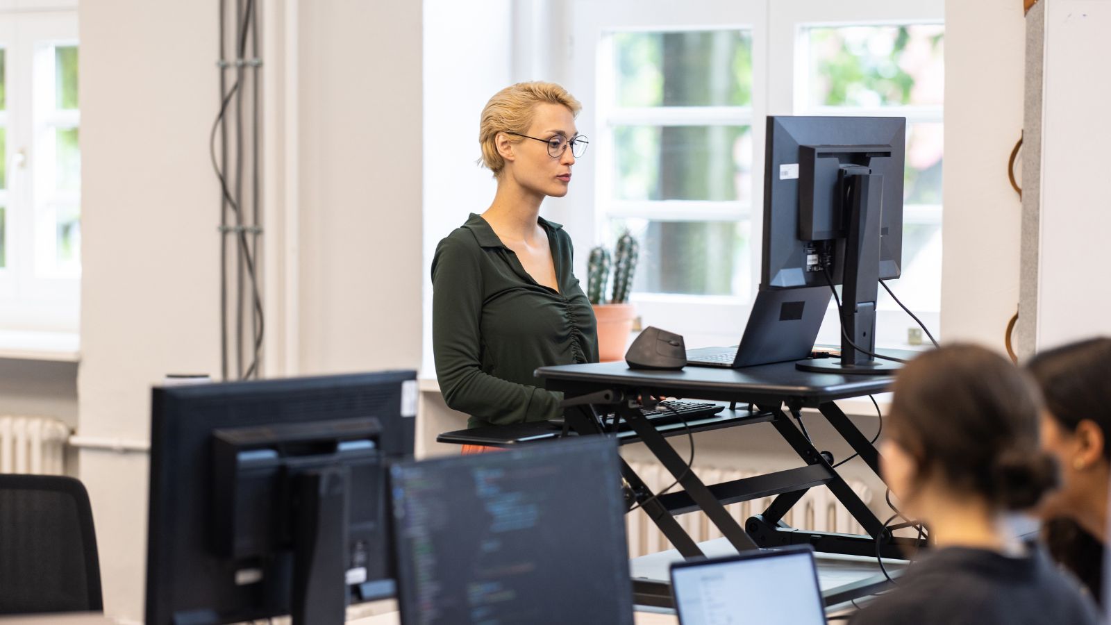 A white person is standing at her ergonomic workstation. Her workstation includes a monitor positioned at eye level.