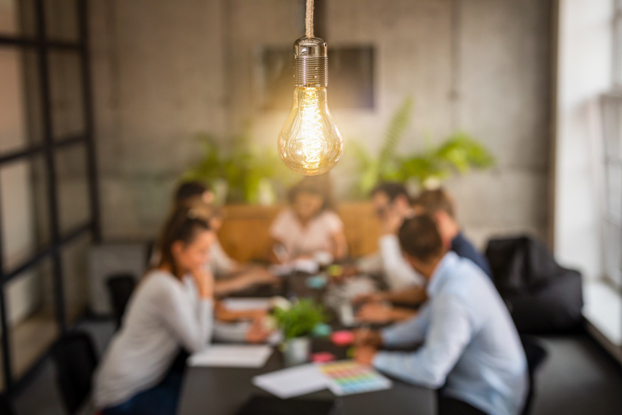 Foreground features a prominent light bulb, while in the background, individuals are engaged in a meeting.