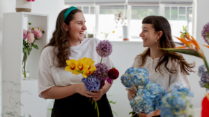 Two people, seemingly co-workers in a flower shop, joyfully looking at each other and laughing, with flowers in their hands.