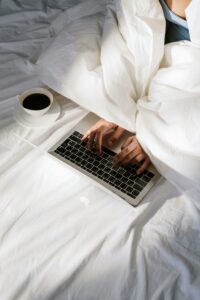 A person working on a laptop while sitting in bed. The bed is covered with a white duvet, and there is a cup of coffee on a saucer next to the laptop.