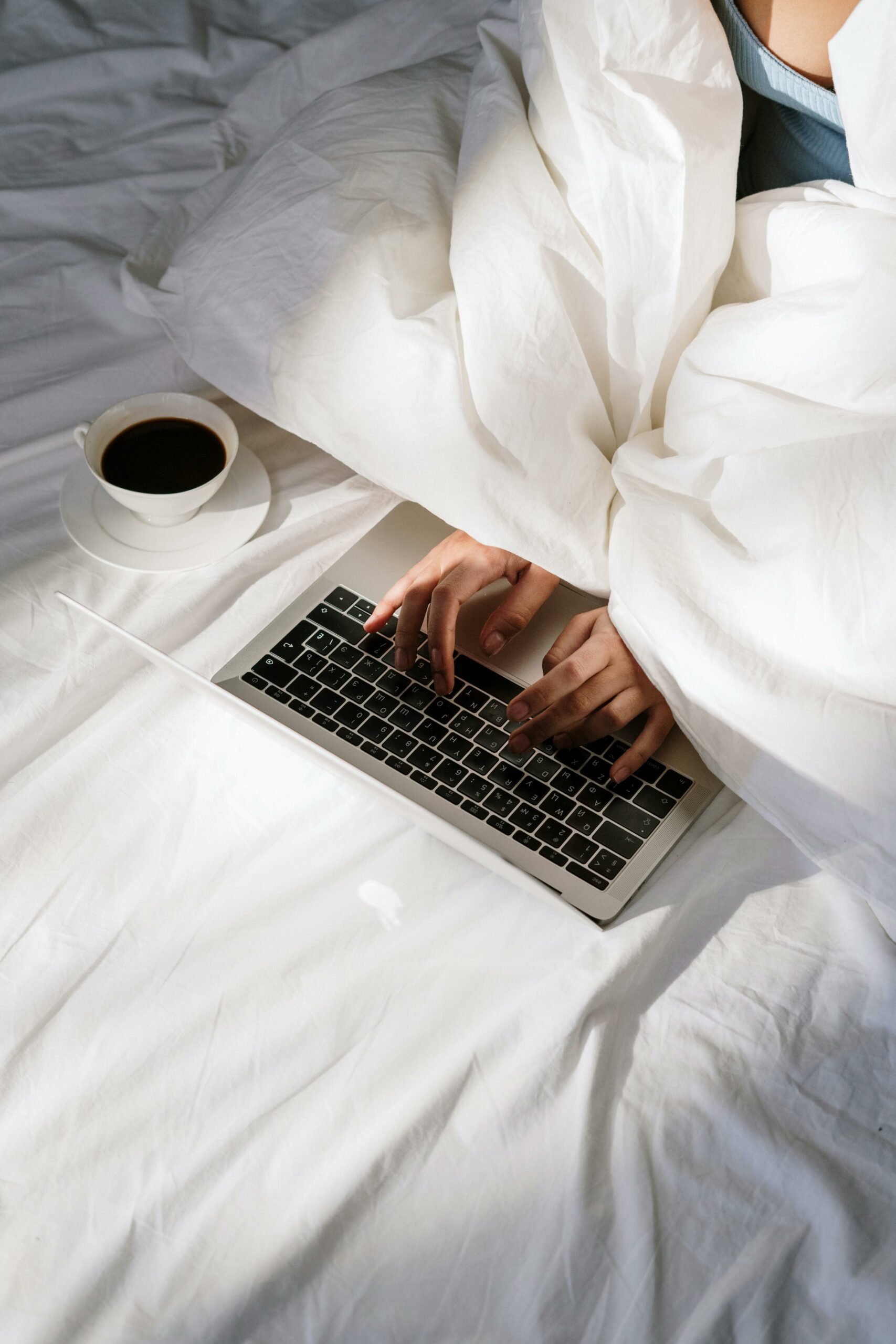 A person working on a laptop while sitting in bed. The bed is covered with a white duvet, and there is a cup of coffee on a saucer next to the laptop.