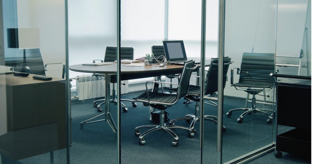 Modern glass-walled conference room with sleek black office chairs on wheels surrounding a circular table. A laptop, papers, and office supplies are neatly arranged on the table, creating a clean and professional atmosphere. The room is well-lit with natural light filtering through the frosted windows.