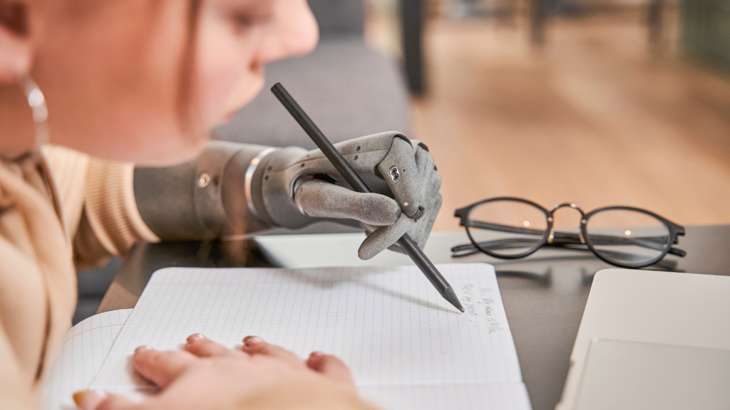 A close-up image shows a person with a prosthetic hand writing in a notebook, highlighting the use of accessibility tools and assistive technology in daily tasks. A pair of glasses rests nearby, symbolizing the integration of devices that support accessibility.