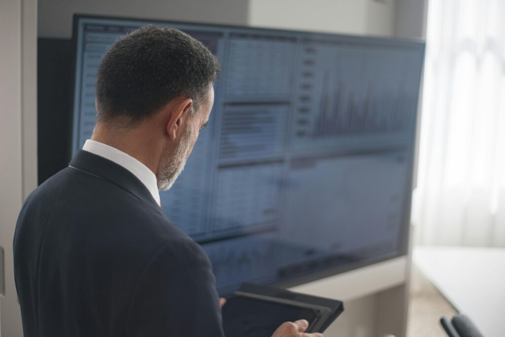 A man in a suit stands in front of a large screen displaying complex data charts, holding a tablet and analyzing information.